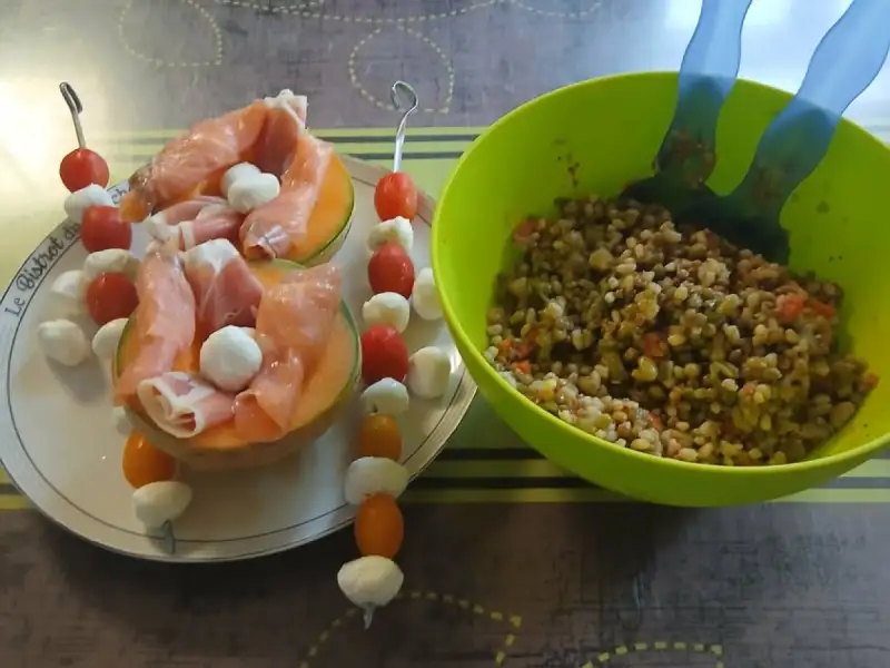 Halbe Melonen mit rohem Schinken und Mozzarellakugeln, Spieße mit Kirschtomaten und Mozzarella, neben einer Salatschüssel